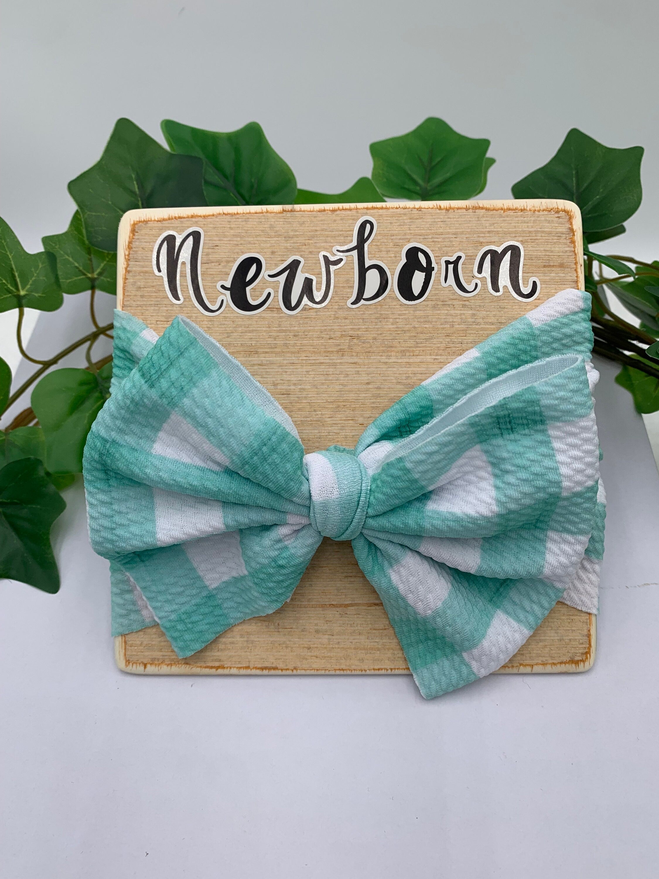 Teal and white checkered bow on a wooden board with 'Newborn' text, surrounded by green leaves.