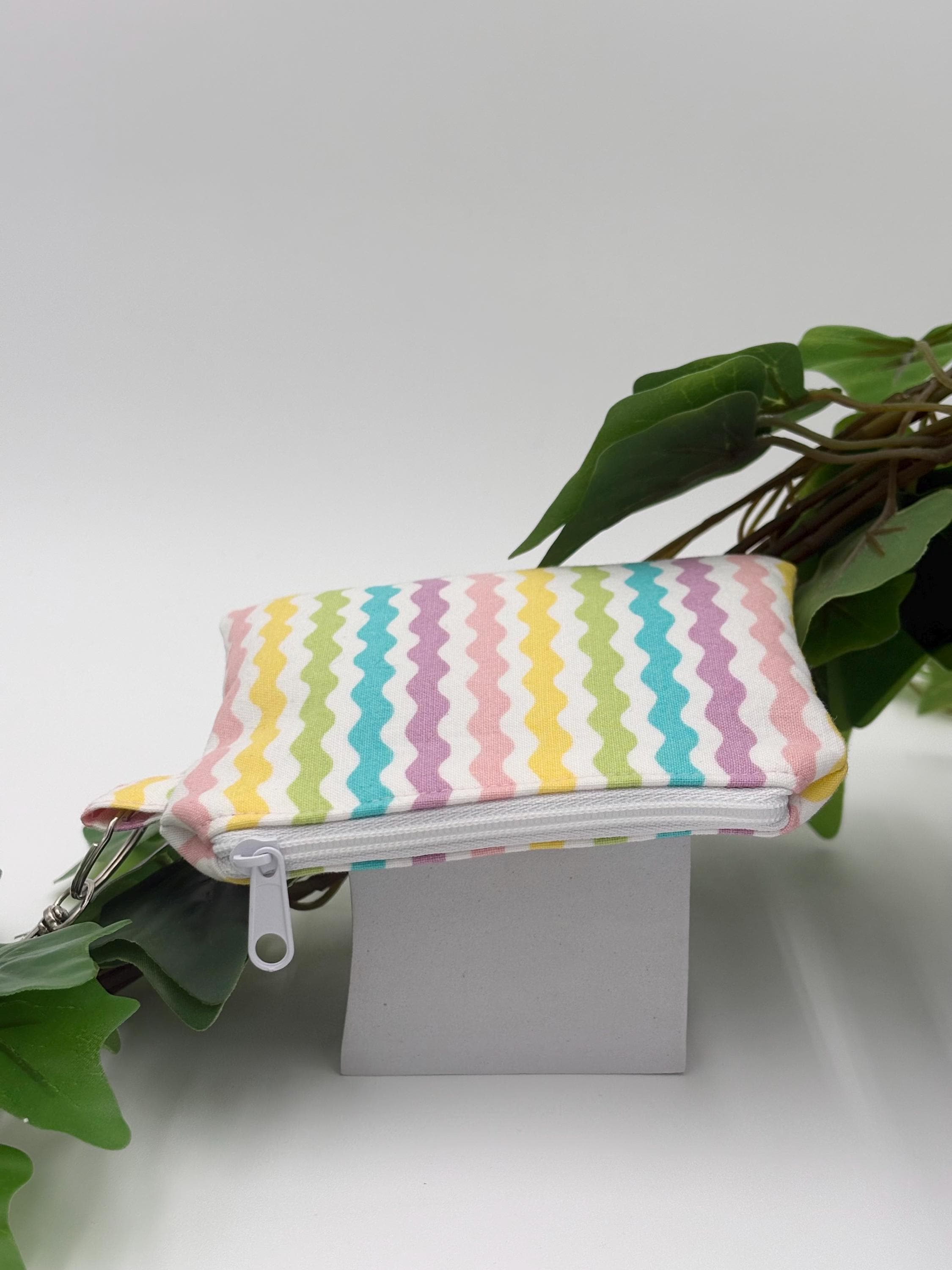 Small coin purse keychain with pastel zigzag pattern with white zipper on a white background with green leaves 