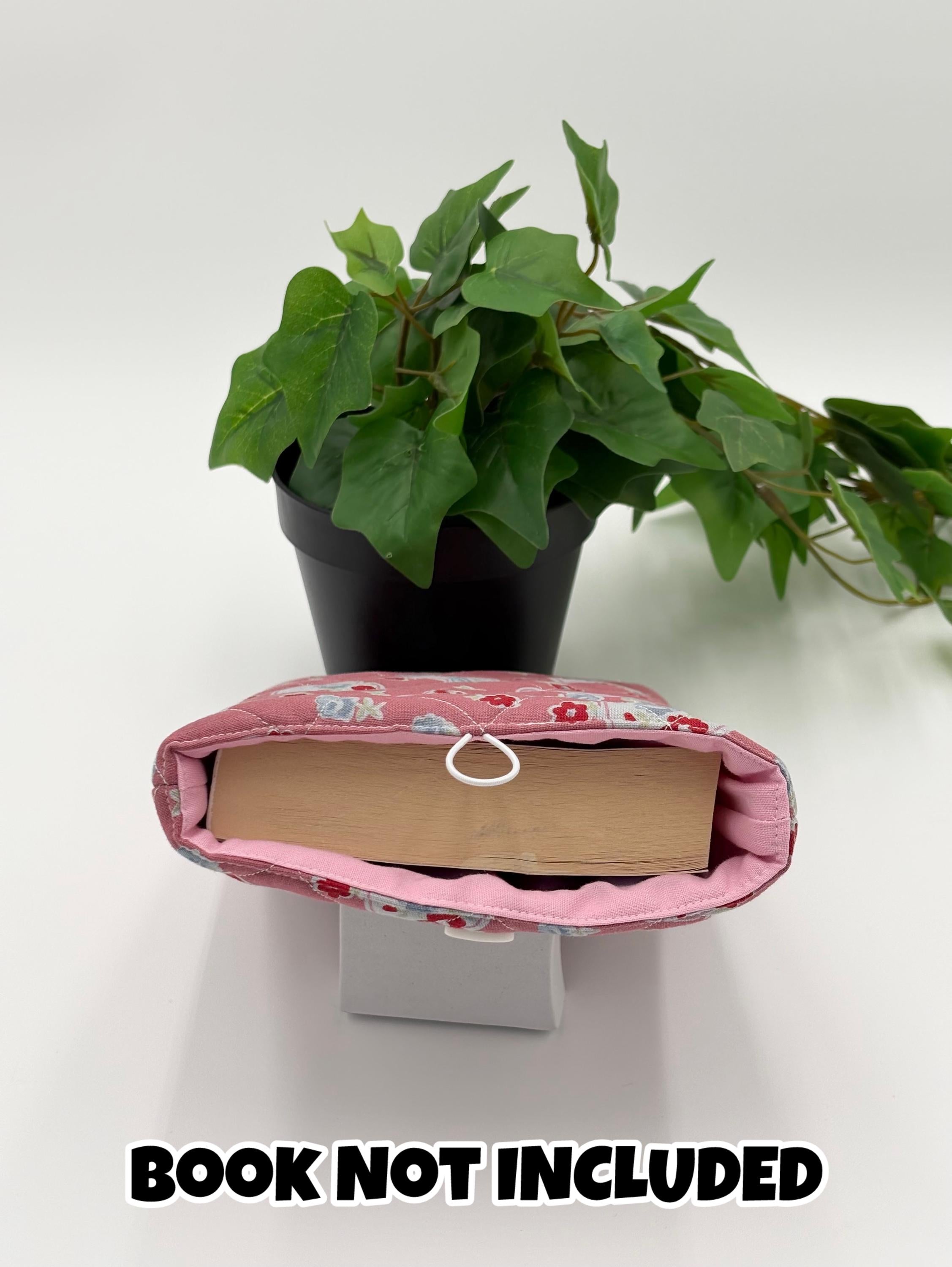 Pink floral and hearts patterned book sleeve with a book inside, placed on a white surface with a green plant in the background with text “book not included”
