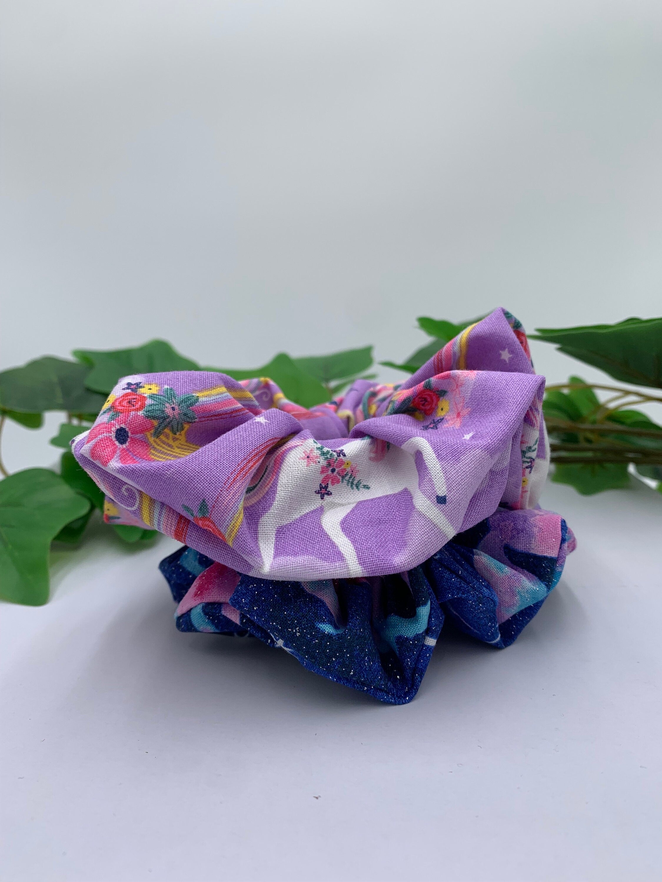 Two colorful scrunchies on a white surface with green leaves in the background