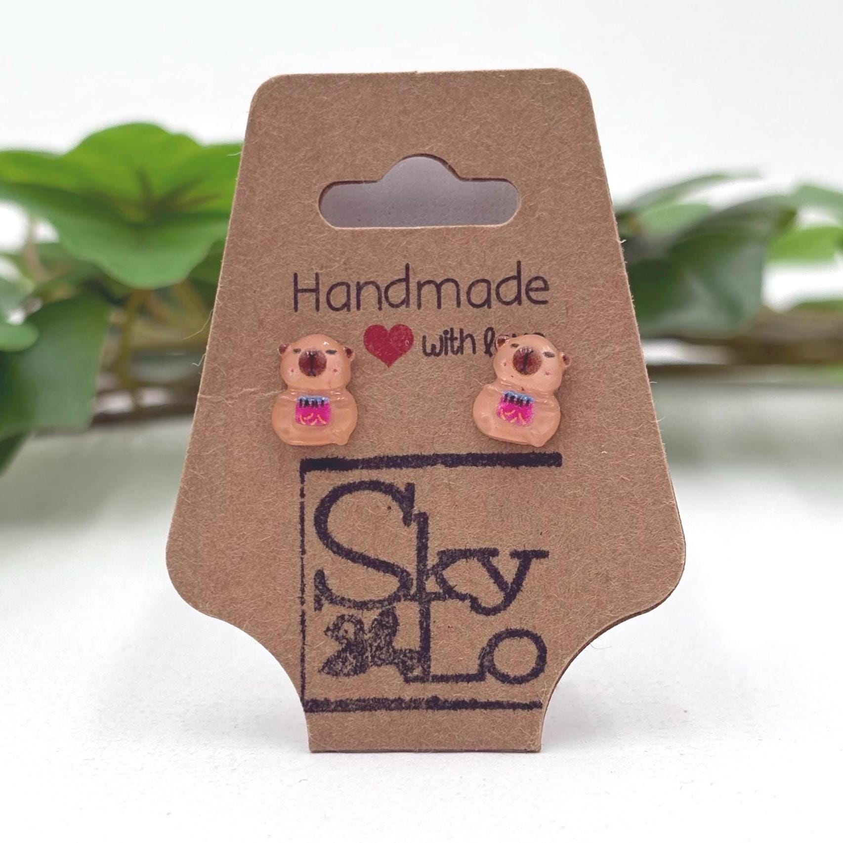  Handmade capybara with a bag of chips earrings on a branded card with a blurred green plant background