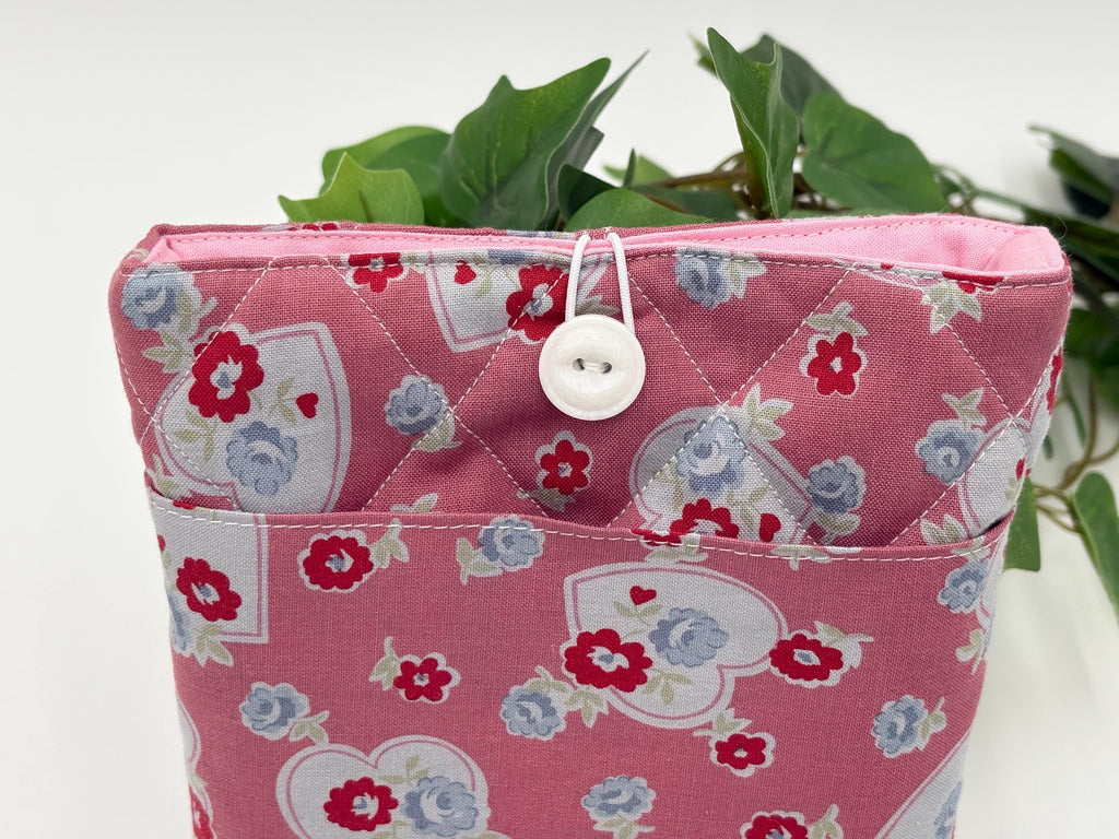 Pink fabric book sleeve with floral and heart pattern and button closure, containing green leaves.