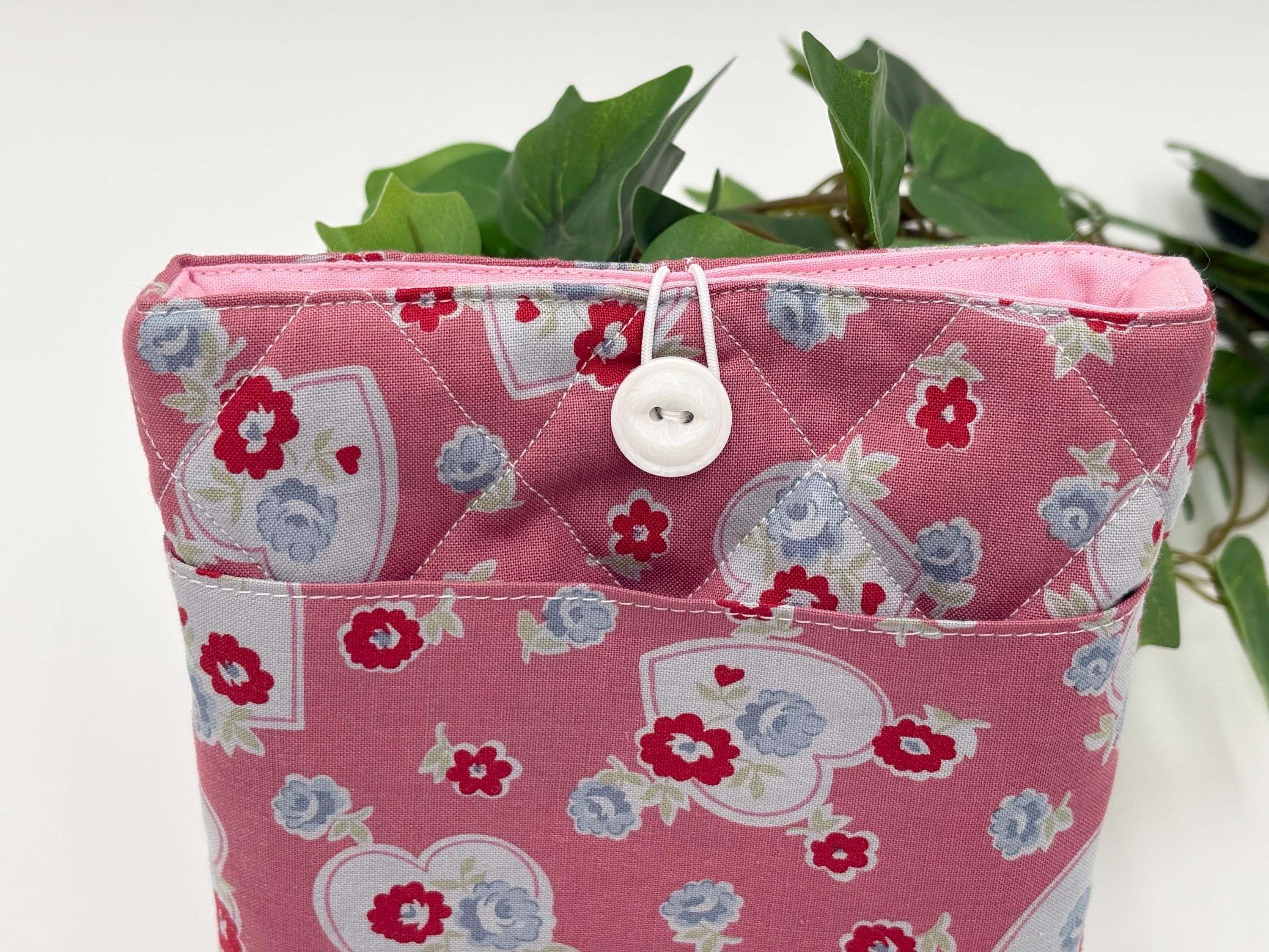 Pink fabric book sleeve with floral and heart pattern and button closure, containing green leaves.