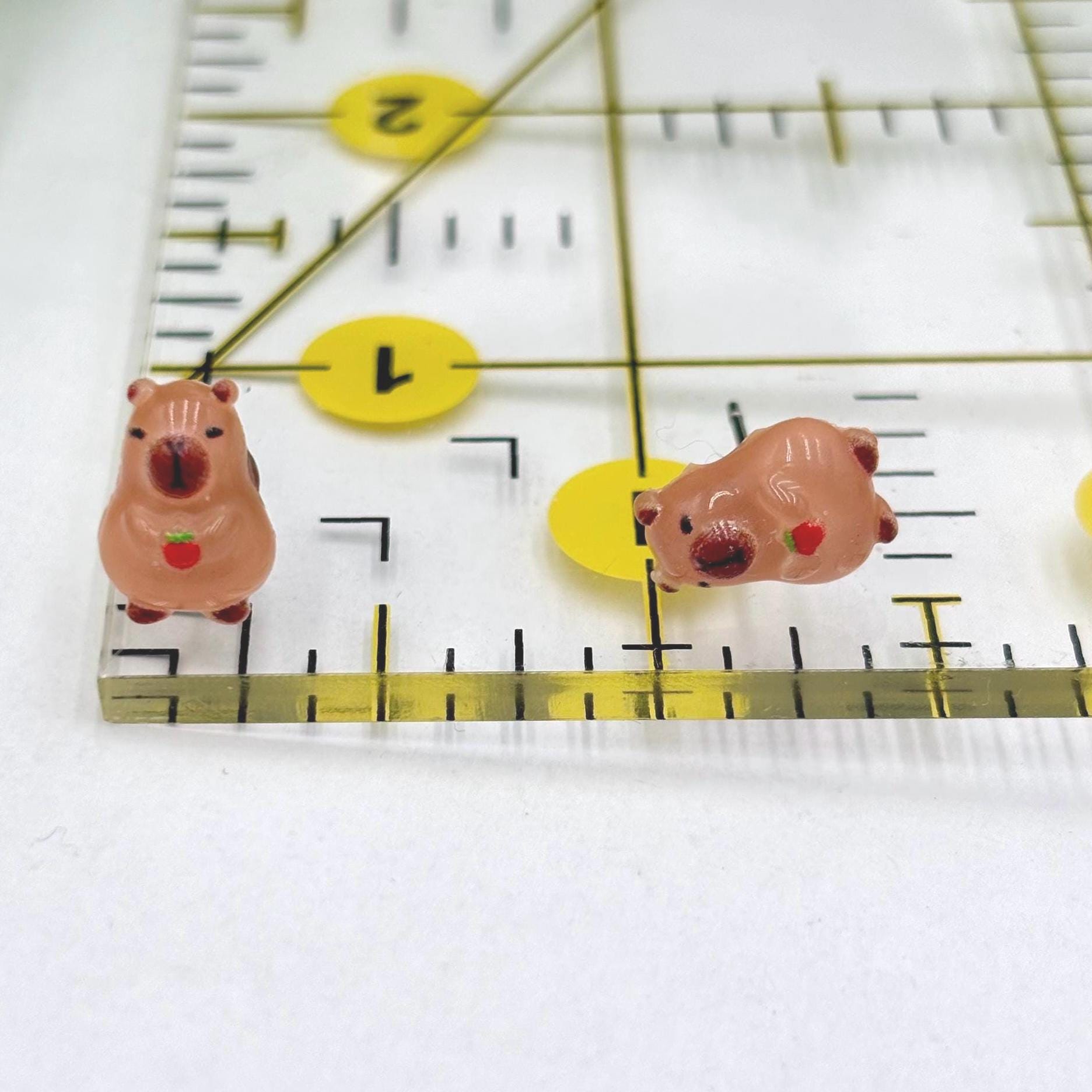 Two capybara with red fruit earrings on a ruler for scale.