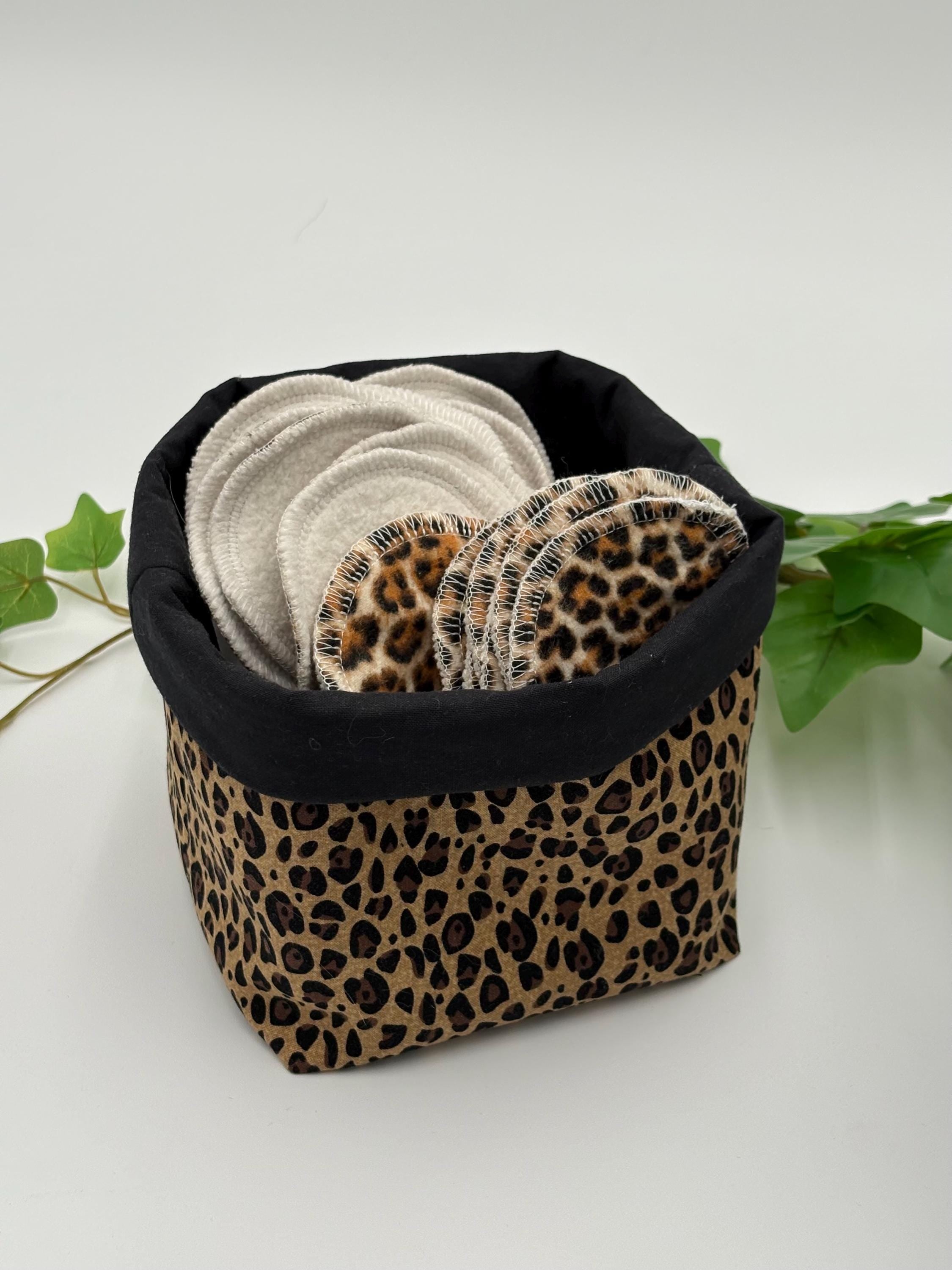 reusable makeup remover pads with leopard print and white fabric in a leopard print and black fabric basket, surrounded by green leaves on a white background.