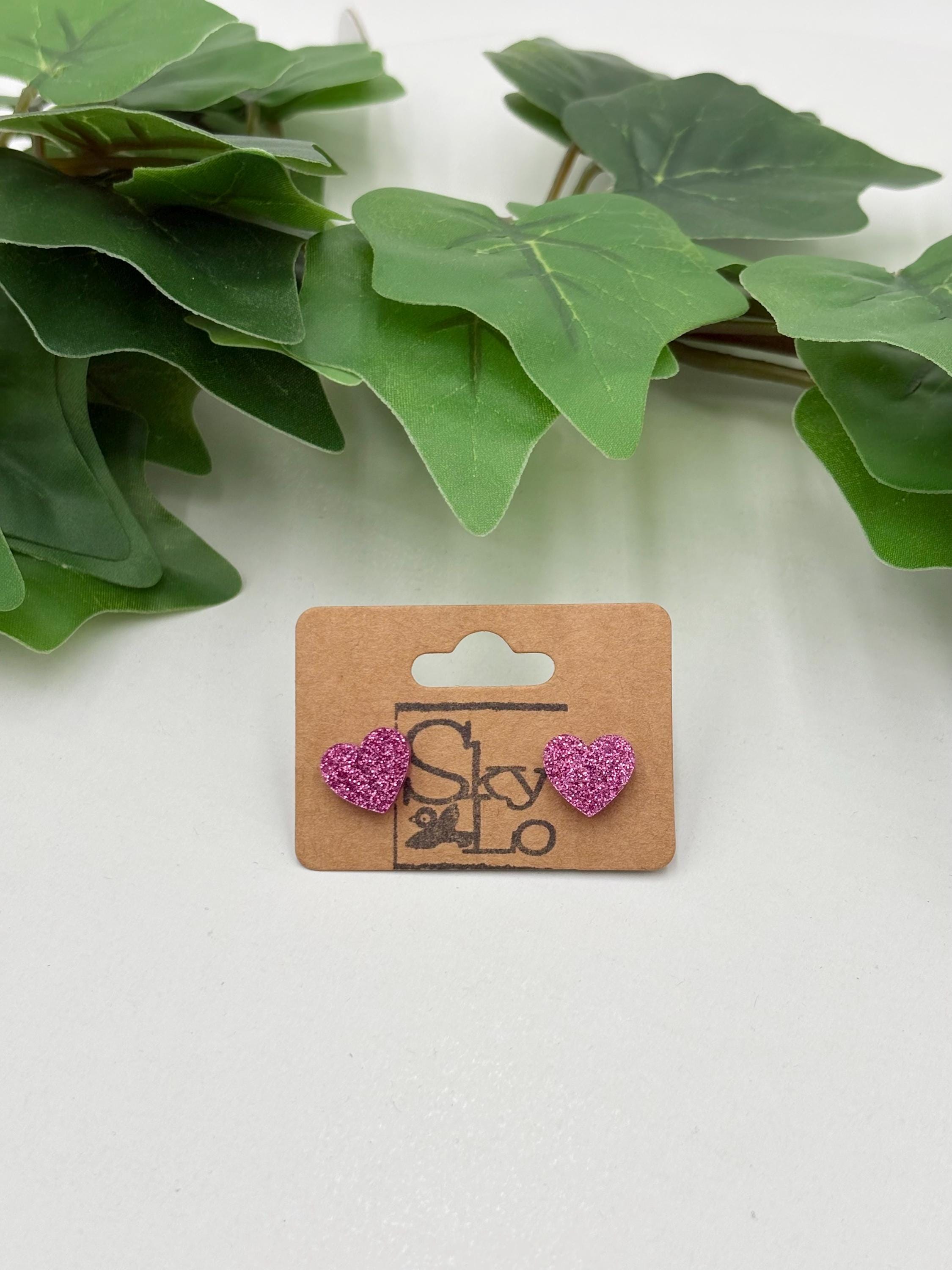 Pink sparkling heart-shaped earrings on a wooden card with a brand logo, placed against green leaves.