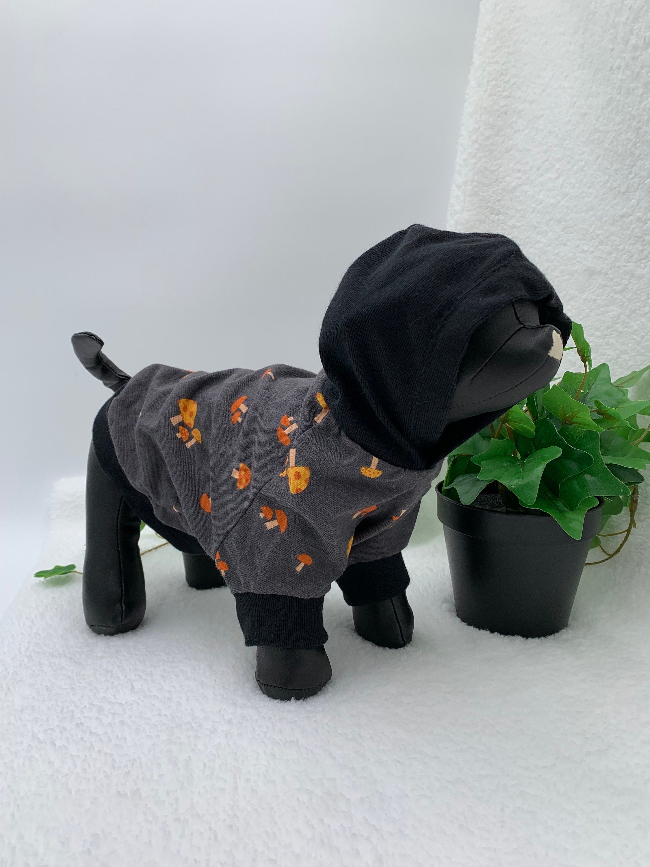 Black dog mannequin wearing a gray mushroom patterned hoodie with hoodie up, standing next to a potted plant on a white surface.