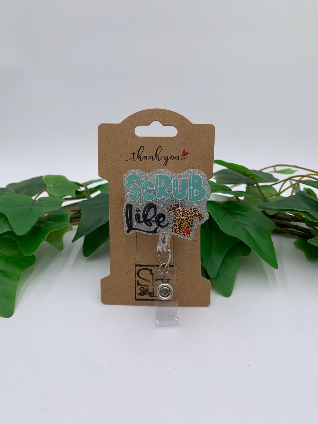 Scrub Life badge holder with decorative charm on a cardboard backing with 'thank you' text, surrounded by green leaves.