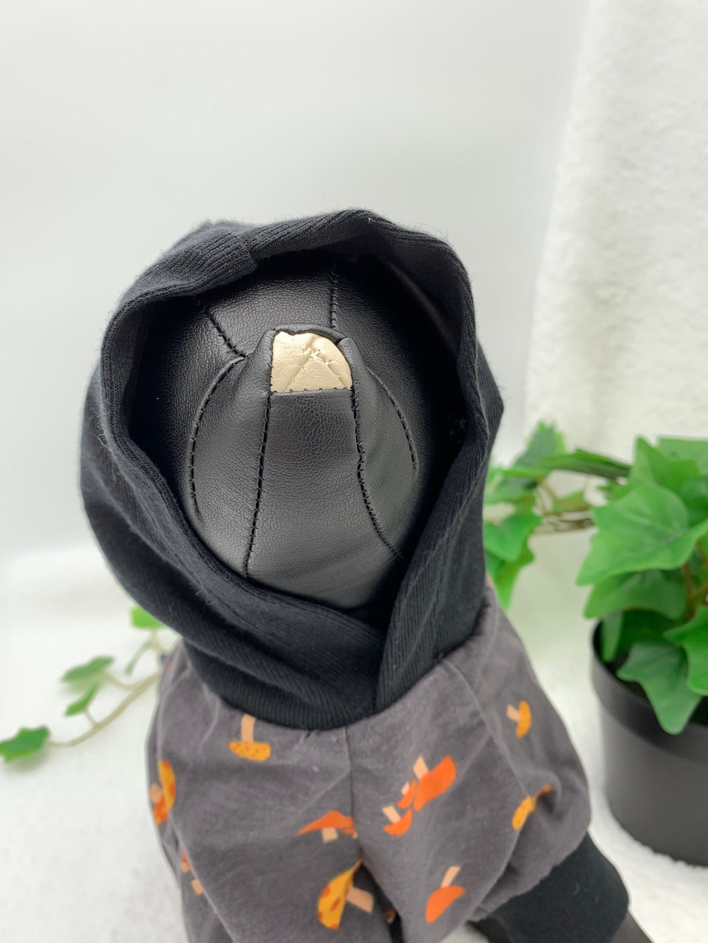 Black dog mannequin wearing a gray mushroom patterned hoodie with hoodie on, standing next to a potted plant on a white surface.
