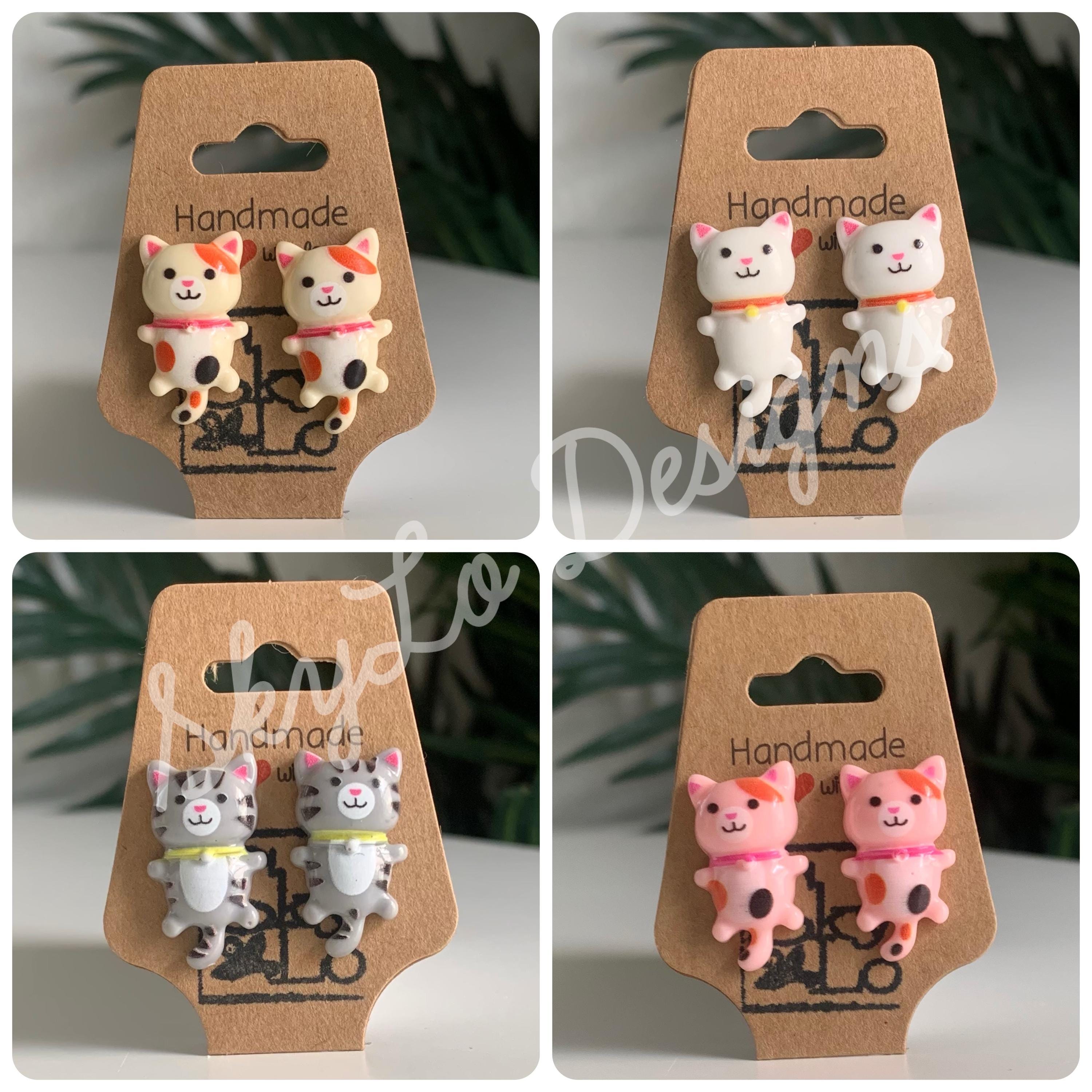 Four pairs of cat-shaped earrings on 'Handmade Designs' cards with a blurred green plant background.