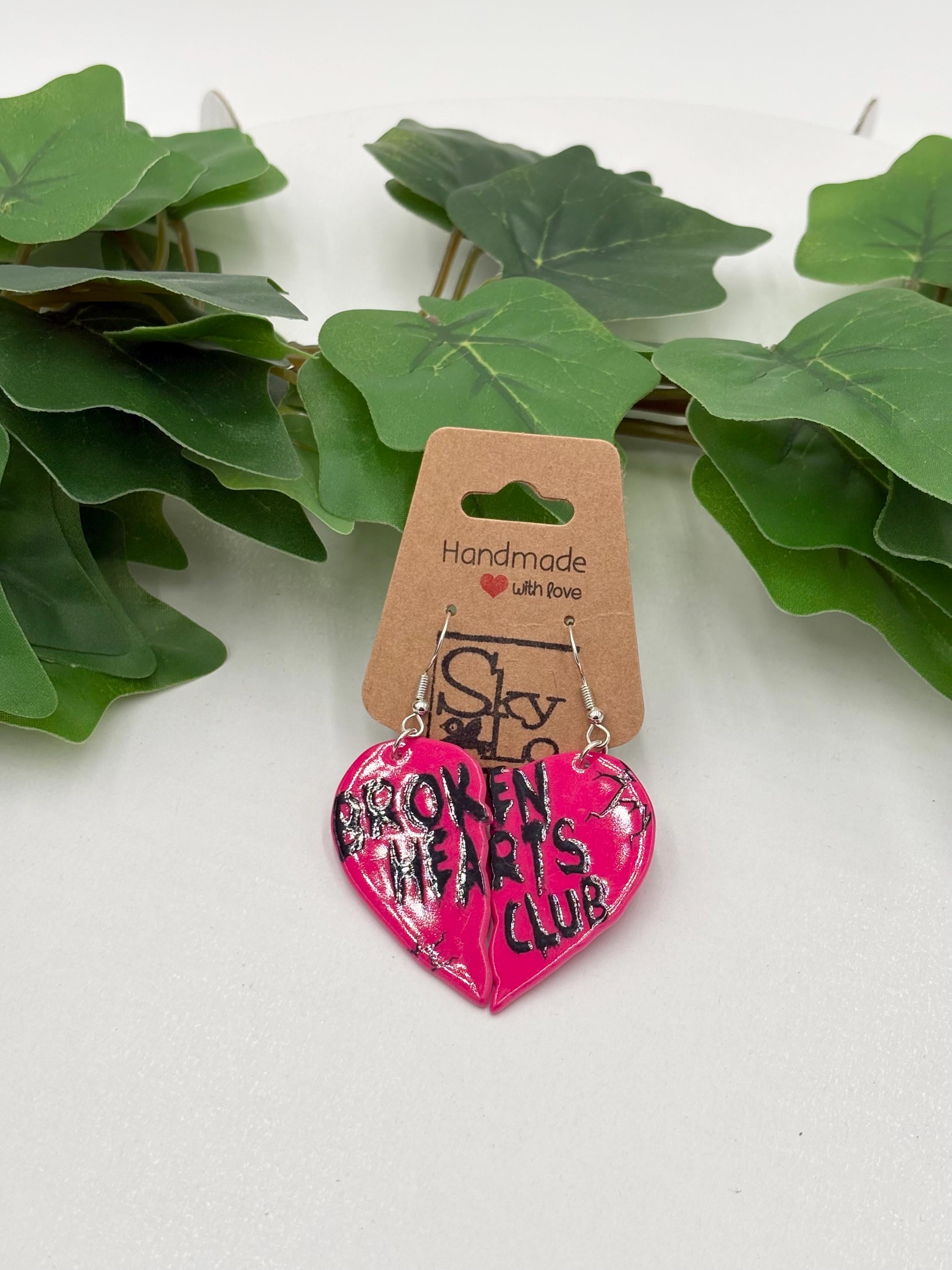 Heart-shaped earrings with 'Broken Hearts Club' text on a white background with green leaves.