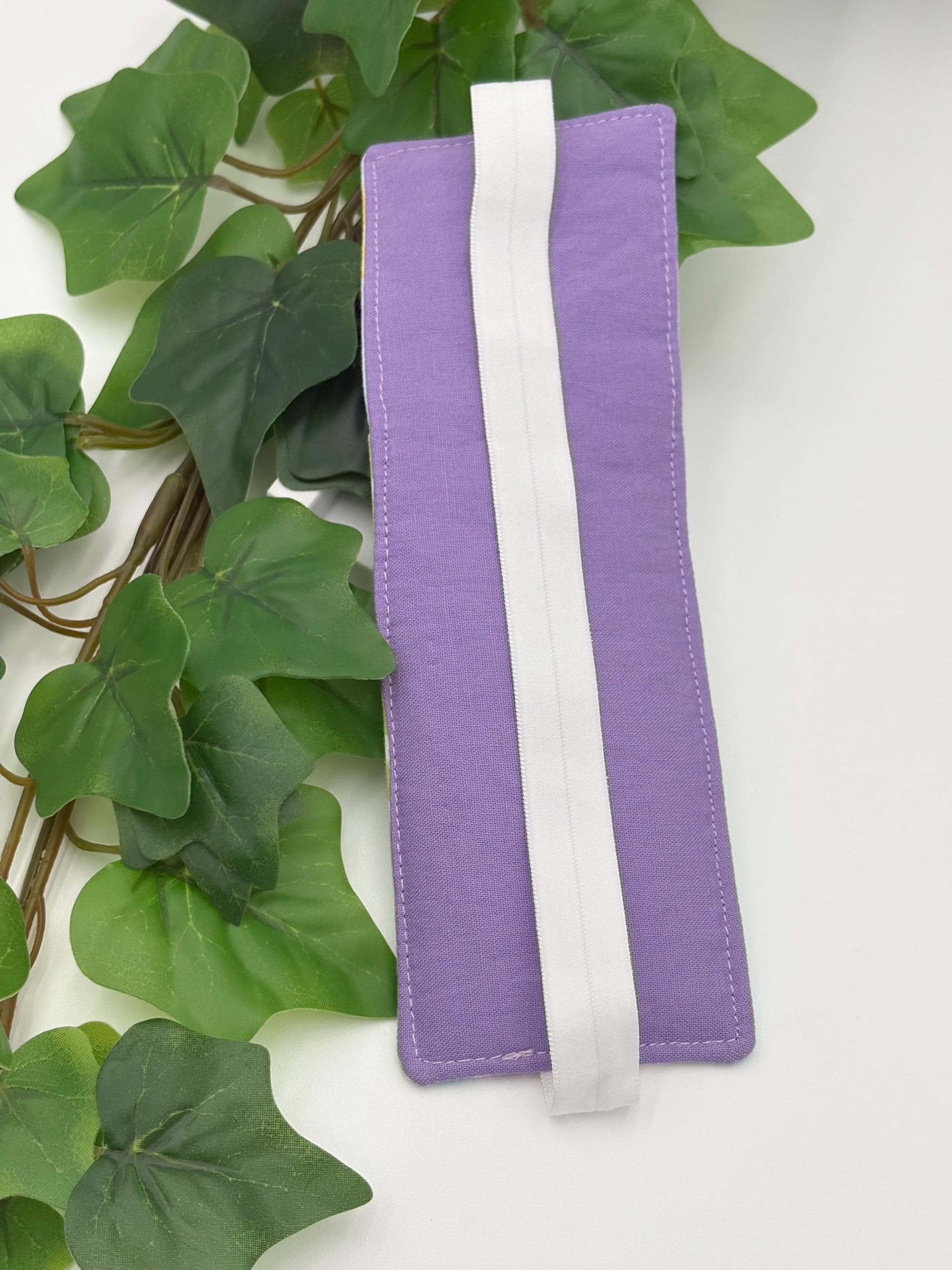 Purple pen holder bookmark with white strap on a white background with green leaves.