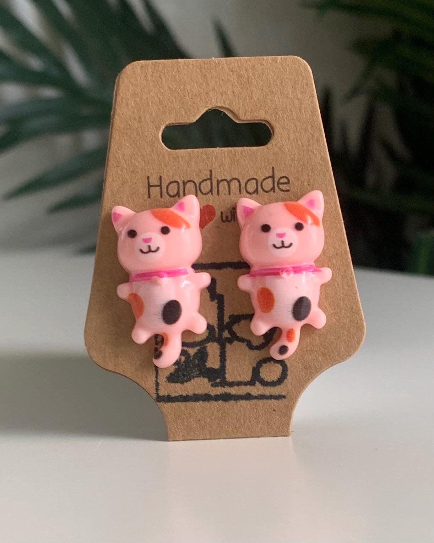Pair of pink calico cat-shaped earrings on a brown card with 'Handmade' text.