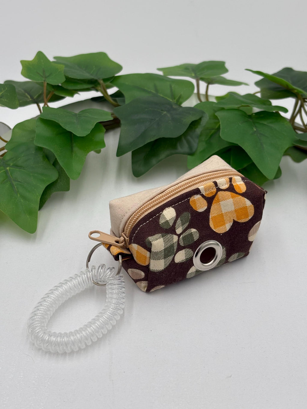 Small fabric dog poop bag holder with paw print design on a white surface with green leaves in the background